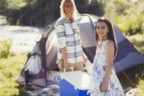 Ritratto madre e figlia sorridente che trasportano il dispositivo di raffreddamento fuori dalla tenda soleggiata del campeggio — Foto stock