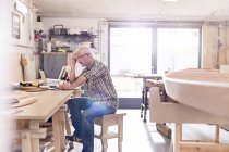 Мужчина плотник, работающий за ноутбуком на рабочем месте возле деревянной лодки в мастерской — стоковое фото
