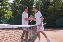 Jovens jogadores de tênis do sexo masculino apertando as mãos no sportsmanship sobre a rede na quadra de tênis de barro ensolarado — Fotografia de Stock