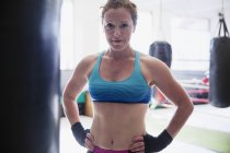Retrato confiante, difícil boxeador feminino em pé no saco de perfuração no ginásio — Fotografia de Stock