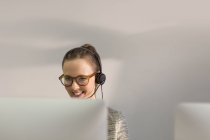 Smiling female telemarketer wearing headset talking on telephone at computer in office — Stock Photo