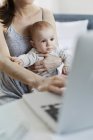 Mãe segurando bebê filha e trabalhando no laptop — Fotografia de Stock