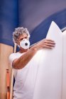 Сосредоточенный мужчина в защитной маске, шлифующий доску для серфинга в мастерской — стоковое фото