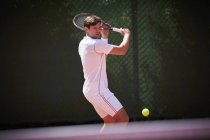 Jovem jogador de tênis masculino jogando tênis, balançando na bola de tênis no campo de tênis ensolarado — Fotografia de Stock