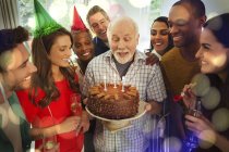 Multiethnische Familie beobachtet Senior beim Ausblasen von Geburtstagskerzen auf Schokoladenkuchen — Stockfoto
