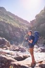 Jeune homme avec sac à dos randonnée et photographie ensoleillé, falaises escarpées avec appareil photo — Photo de stock