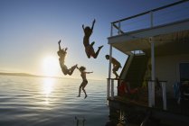 Jóvenes amigos adultos saltando de la casa flotante de verano en el océano puesta de sol - foto de stock