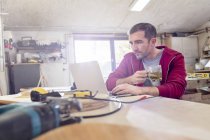 Hombre carpintero beber té y trabajar en el ordenador portátil en el banco de trabajo en el taller - foto de stock