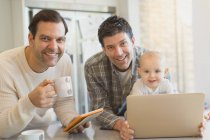 Portrait smiling male gay parents with baby son using digital tablet and laptop in kitchen — Stock Photo
