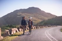 Young couple with backpacks hiking along remote, sunny road — Stock Photo