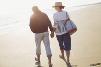 Barefoot mature couple holding hands and walking on sunny beach — Stock Photo