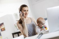 Mother holding baby daughter and working at laptop and talking on cell phone — Stock Photo
