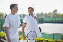 Smiling young male tennis players walking with tennis rackets along sunny tennis court — Stock Photo