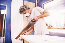 Male surfboard designer wearing protective mask and using saw in workshop — Stock Photo