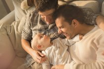 Vista de acima masculino gay pais abraçando com bonito bebê filho no sofá — Fotografia de Stock