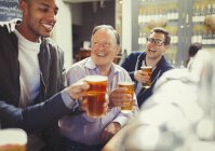 Hombres sonrientes amigos brindando vasos de cerveza en el bar - foto de stock