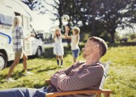 Sereno padre che riposa sulla poltrona con la famiglia che gioca in background al di fuori del camper soleggiato — Foto stock