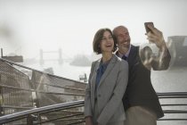 Улыбчивая деловая пара делает селфи с телефоном на солнечном городском мосту, Лондон, Великобритания — стоковое фото