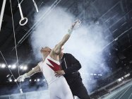 Тренер поднимает мужскую гимнастку ниже колец гимнастики на арене — стоковое фото