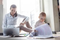 Curious baby daughter looking at paperwork next to mother working at laptop — Stock Photo