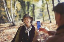 Mujer mayor juguetona siendo fotografiada por el marido con el teléfono de la cámara en el parque de otoño - foto de stock