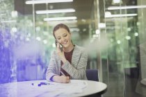 Улыбающаяся деловая женщина разговаривает по мобильному телефону и делает заметки в конференц-зале — стоковое фото