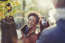Femme souriante posant pour petit ami avec téléphone caméra dans les bois ensoleillés d'automne — Photo de stock