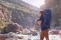 Young man with backpack hiking photographing with camera below sunny, remote cliffs — Stock Photo