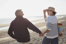 Смеющиеся зрелые пары держатся за руки и ходят по солнечному пляжу — стоковое фото
