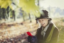 Smiling senior woman using cell phone in sunny autumn park — Stock Photo