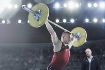 Trainer beobachtet männlichen Gewichtheber, wie er Langhantel über Kopf in Arena hockt — Stockfoto