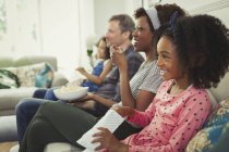 Joven familia multiétnica viendo películas y comiendo palomitas en el sofá - foto de stock