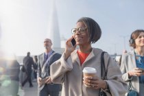 Smiling businesswoman drinking coffee and talking on cell phone on sunny busy urban pedestrian bridge — Stock Photo