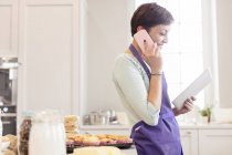 Fêmea cozimento caterer, falando no telefone celular e usando tablet digital na cozinha — Fotografia de Stock