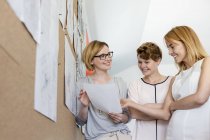 Sorrindo designers femininos revisando provas no quadro de avisos no escritório — Fotografia de Stock