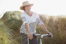 Усміхнена зріла жінка їде на велосипеді на сонячному пляжі трава — стокове фото
