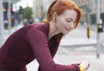 Усміхнена жінка бігунка з червоним волоссям і навушниками перевіряє розумний годинник — стокове фото