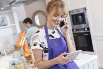 Усміхнений доглядач працює, розмовляє на мобільному телефоні та використовує цифровий планшет на кухні — стокове фото