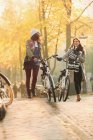 Giovani amiche a piedi biciclette lungo la soleggiata strada autunnale — Foto stock
