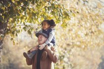 Grandfather carrying daughter on shoulders below trees in sunny autumn park — Stock Photo
