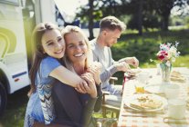 Smiling, affectionate mother and daughter hugging at table outside sunny motor home — Stock Photo