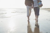 Affectionate barefoot mature couple walking, holding hands in sunny ocean beach surf — Stock Photo
