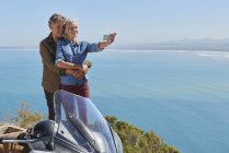 Старша пара бере селфі поруч з мотоциклом з видом на сонячний океан — стокове фото