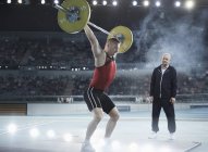 Trainer beobachtet männlichen Gewichtheber, wie er Langhantel über Kopf in Arena hockt — Stockfoto