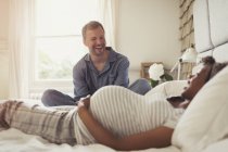Багатоетнічна вагітна пара розслабляється і сміється на ліжку — стокове фото