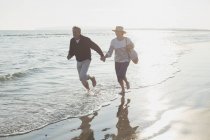Playful mature couple holding hands and running in sunny ocean surf — Stock Photo