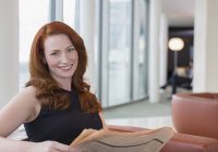 Portrait smiling businesswoman with red hair reading newspaper in office lounge — Stock Photo