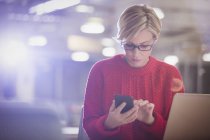 Femme d'affaires travaillant tard à l'ordinateur portable, textos avec téléphone portable dans le bureau sombre — Photo de stock