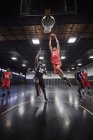 Jovem jogador de basquete masculino pulando para bater basquete afundado em jogo na quadra no ginásio — Fotografia de Stock