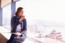 Empresária pensativa com tablet digital bebendo café na janela urbana ensolarada — Fotografia de Stock